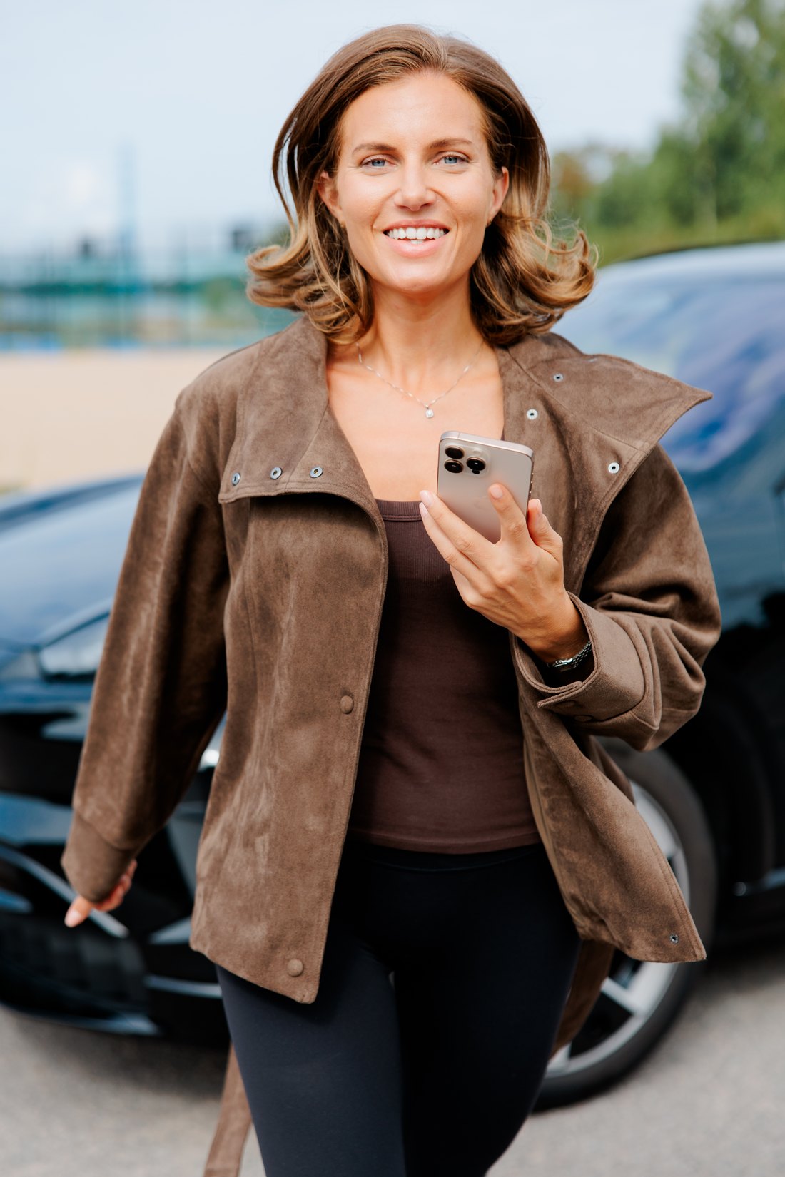 Jenna Toivakka looking at the camera, phone in hand, a dark car parked behind her.