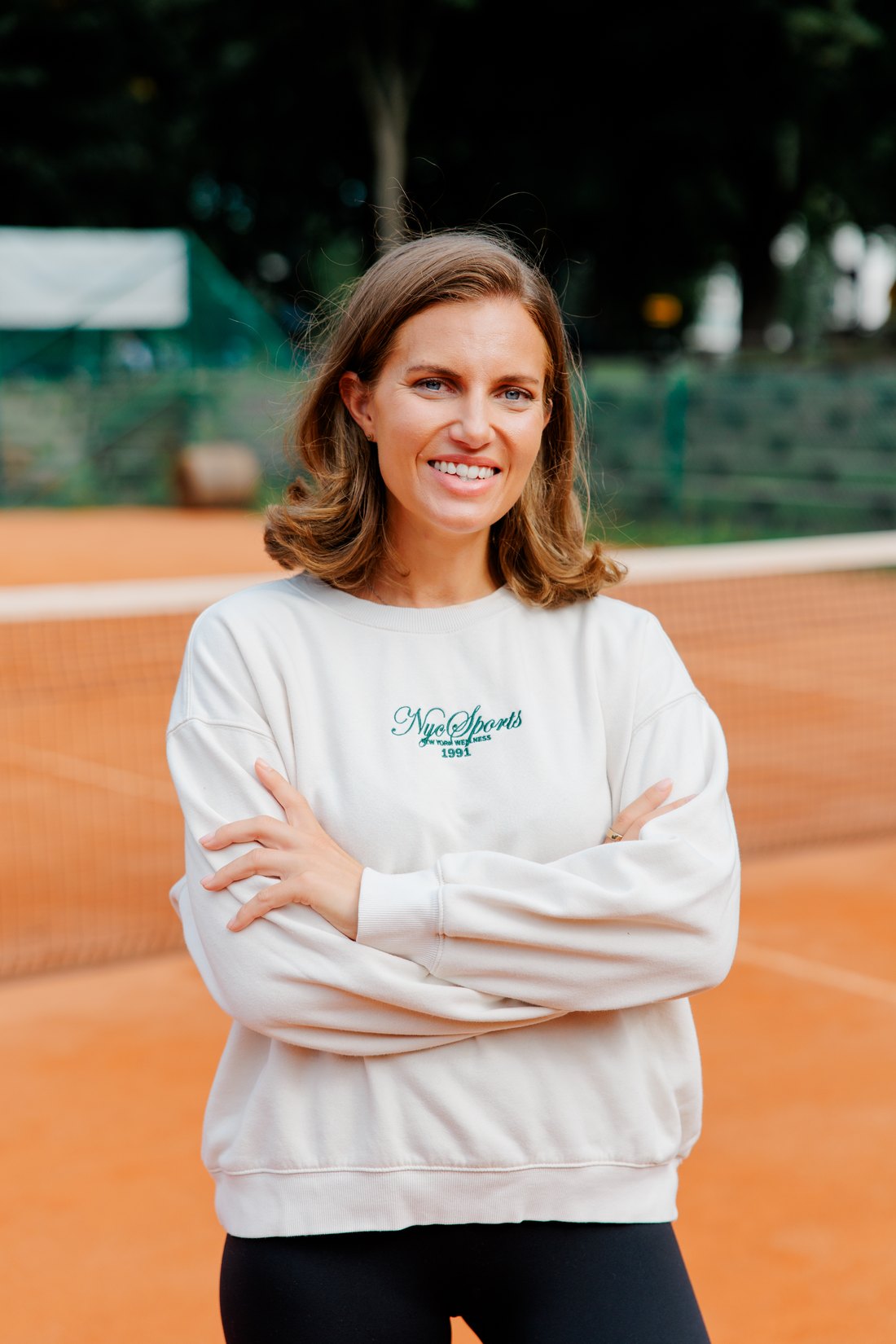 Jenna Toivakka on a clay tennis court, arms folded, in a cream sweatshirt.